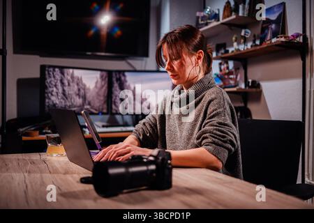 Una donna con maglione grigio si concentra sul proprio laptop a una scrivania con fotocamera, bevande e due monitor in un ambiente caldo e creativo in casa. Foto Stock
