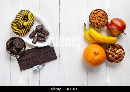 Confronto tra scelte alimentari sane e malsane. Vista dall'alto di frutta e noci sani in contrasto con cioccolato zuccherino, biscotti e ciambelle su un bianco Foto Stock