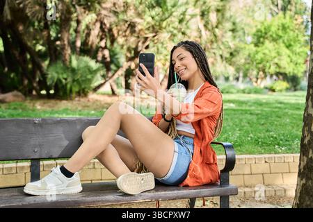 La giovane donna alla moda con lunghe trecce si sta facendo un selfie mentre si gusta una bevanda fresca sulla panchina del parco, godendosi una rilassante giornata estiva Foto Stock