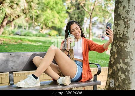 Una giovane donna con lunghe trecce si gode un rinfrescante succo verde mentre si fa un selfie sulla panchina del parco Foto Stock