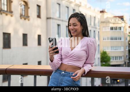 Giovane donna felice con lunghe trecce appoggiate su una ringhiera del balcone, che utilizza il suo smartphone e sorride in un ambiente soleggiato della città Foto Stock