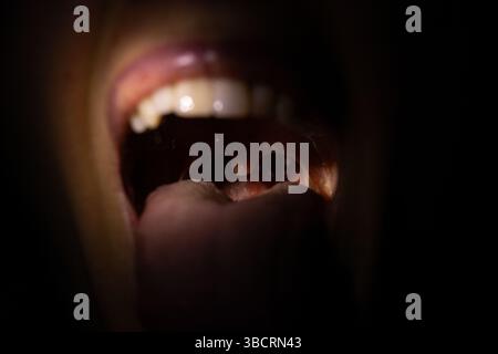 Bocca aperta di una ragazza al buio con un fascio di luce in bocca con lingua e denti visibili, bocca larga aperta Foto Stock