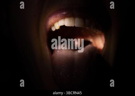 Bocca aperta di una ragazza al buio con un fascio di luce in bocca con lingua e denti visibili, bocca larga aperta Foto Stock