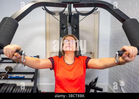Donna matura che fa esercizi di bodybuilding per il petto con una macchina per il peso a casa. Foto Stock
