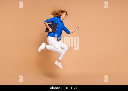Giovane donna d'affari sorridente che salta energicamente con un notebook in mano su uno sfondo beige, mostrando stile ed entusiasmo Foto Stock