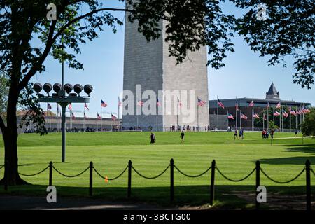 Washington Monument sul National Mall di Washington DC | WASHINGTON D.C., Stati Uniti: I visitatori si riuniscono alla base del Washington Monument sul National Mall, circondato da file di bandiere americane. L'obelisco in marmo e granito, completato nel 1884, onora George Washington e si erge a 555 piedi 5⅛ pollici (169,29 m), rendendolo la struttura in muratura più alta del mondo. Cinquanta palafitte in alluminio, installate nel 1959, circondano la base del monumento. Foto Stock