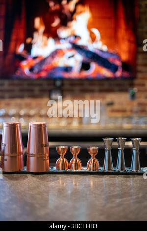 Attrezzatura da bar: bicchieri di rame e misurini sul bancone | Bicchieri di rame e misurini di metallo disposti su un bancone da bar, pronti per la preparazione di cocktail. I recipienti di rame lucidati, probabilmente progettati per bevande come i Moscow Mule, contrastano con gli strumenti di misurazione in acciaio inossidabile essenziali per una mixologia precisa. Questa collezione di attrezzature professionali da bar rappresenta gli strumenti fondamentali necessari per creare cocktail coerenti e ben bilanciati in un ambiente da bar. Foto Stock