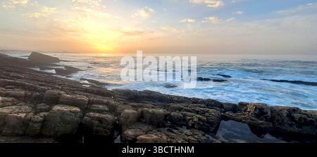 L'alba dorata dipinge il cielo su una costa rocciosa mentre le onde si infrangono contro la riva, creando un paesaggio marino pittoresco Foto Stock