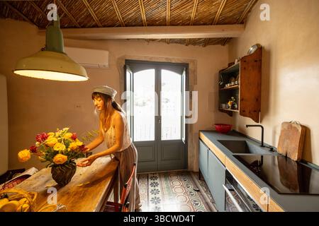 Donna che organizza fiori freschi in una cucina rustica illuminata dal sole con design tradizionale siciliano, piastrelle a motivi geometrici e una vista della storica città collinare attraverso doppie porte aperte Foto Stock