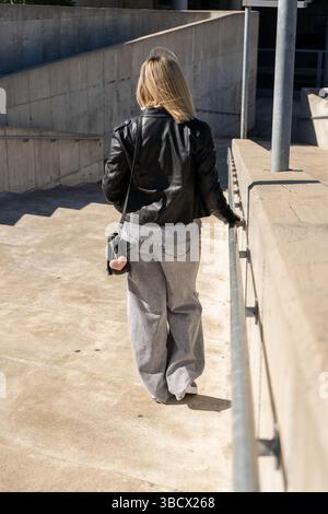 Donna bionda che indossa una giacca in pelle nera e jeans grigi che scendono giù per le scale di cemento in un ambiente urbano Foto Stock