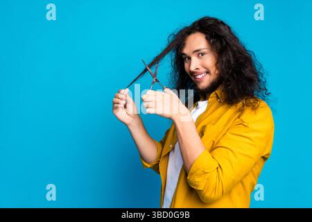 Un giovane sicuro rifinisce i suoi lunghi capelli ricci con delle forbici su uno sfondo blu brillante, sorridendo allegramente Foto Stock