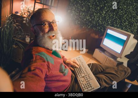 Uomo anziano sorridente mentre lavora su un computer a casa in un accogliente spazio ufficio tra piante verdi Foto Stock