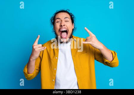 Ritratto di un uomo entusiasta con una camicia gialla che fa gesti di roccia su un vivace sfondo blu Foto Stock