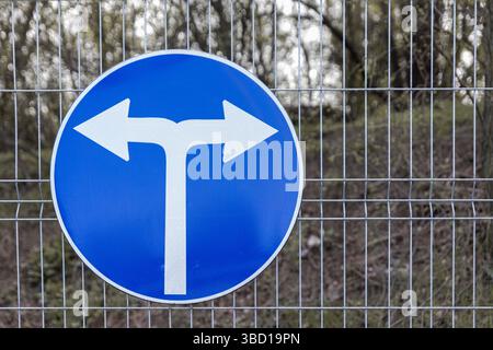 Segnaletica stradale circolare blu che indica una svolta obbligatoria a sinistra o a destra, montata su una recinzione metallica con un'area boscosa sullo sfondo. Foto Stock