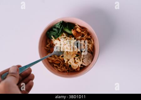 Un pasto di noodle salato è tenuto da una mano, pronto per essere consumato. Il piatto include polpette e verdure verdi. Foto Stock