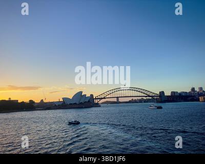 L'Opera House di Sydney e l'Harbour Bridge al tramonto, con barche che navigano attraverso l'acqua scintillante sotto un cielo sereno. Foto Stock