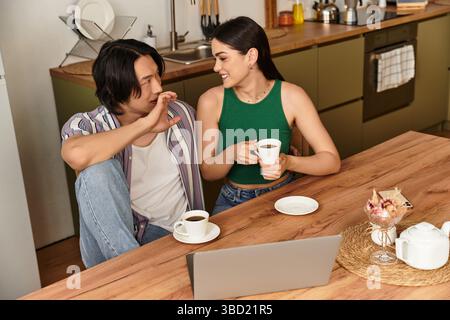 La coppia si siede vicino a un tavolo di legno, condividendo risate e bevande calde in una cucina luminosa. Foto Stock