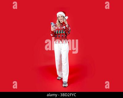 Affascinante Santa donna caucasica che tiene il telefono. Bionda sorridente che guarda e chatta sullo smartphone su sfondo rosso isolato. Natale. Foto Stock