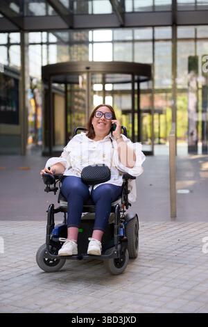 Donna d'affari sorridente in sedia a rotelle che fa una telefonata fuori dall'edificio degli uffici - concetto di sedia a rotelle e disabilità Foto Stock