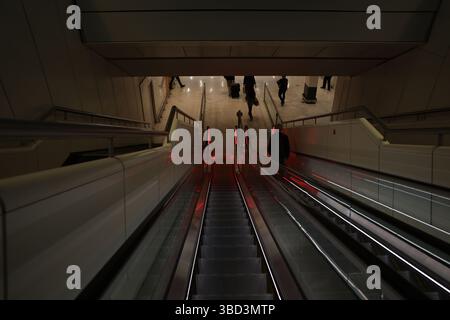 Una scala mobile elegante e moderna scende in un livello inferiore di uno spazio pubblico, fiancheggiata da pannelli di vetro e illuminata da luci rosse. Una grossa scre Foto Stock