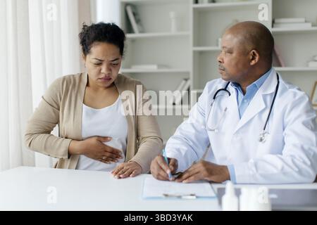 Paziente di colore che tiene lo stomaco e soffre di dolore addominale durante l'incontro con il medico Foto Stock