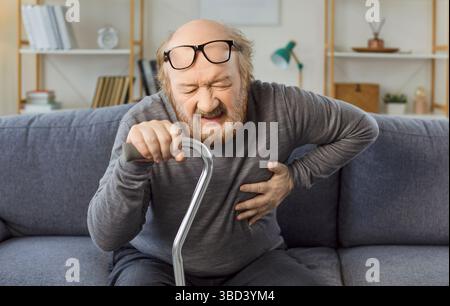 Uomo anziano seduto sul divano con la canna, che stringe il petto nel dolore e soffre di infarto. Foto Stock