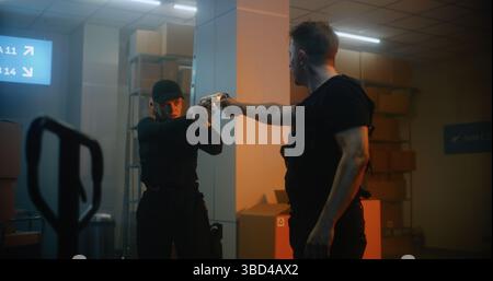 Armed Confrontation: Uomo e donna in Tactical Gear che puntano le pistole l'uno contro l'altro e parlano. Ufficiale che attacca Criminal in Dark Warehouse o Storage. Standoff teso e scena drammatica di Action Movie. Foto Stock