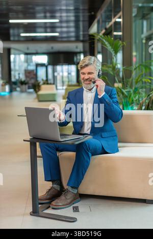 Uomo d'affari caucasico che indossa cuffie, call center o assistenza telefonica, parla con il cliente o con il collega nell'atrio dell'ufficio. Un ragazzo in tuta si siede sul divano dell'hotel. Foto Stock