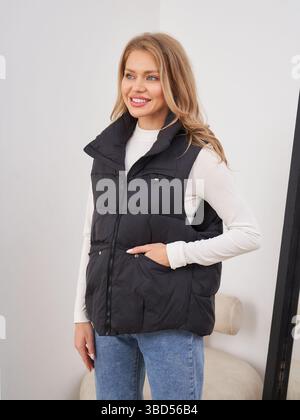 Donna sorridente con camicia bianca a manica lunga e gilet nero con piumino, abbinata a jeans blu. Le sue mani riposano nelle tasche del giubbotto. Capelli biondi lunghi alle spalle Foto Stock