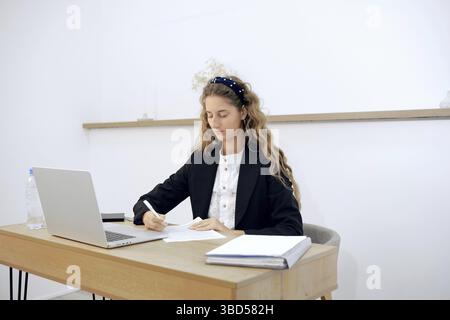 In un ufficio luminoso, una giovane donna si concentra sulla scrittura di appunti alla scrivania mentre un notebook è aperto nelle vicinanze. Una bottiglia d'acqua si siede accanto a lei mentre si prepara Foto Stock