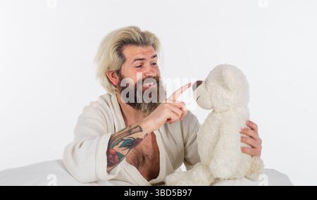 Uomo con orsacchiotto. Uomo barbuto che riposa a letto con orsacchiotto. L'hipster sorridente in accappatoio gioca con un simpatico giocattolo di orsacchiotto. Giocattolo morbido per dormire Foto Stock