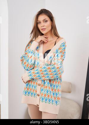 Donna con un cardigan colorato sopra la parte superiore nera, che si piega lateralmente con la mano che tocca il mento, guardando verso la fotocamera. Sedia dai toni neutri Foto Stock
