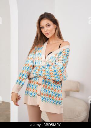 Donna che indossa un cardigan colorato, in piedi in una stanza con arco e sedia; cardigan leggermente distaccato, che mostra sicurezza e. Foto Stock