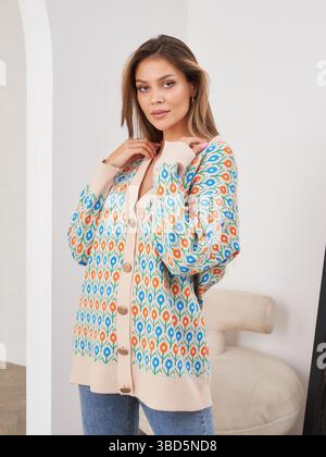 Elegante donna con cardigan e jeans dai motivi colorati, è in piedi con sicurezza all'interno, espressione neutra sul viso, atmosfera accogliente con morbida sedia Foto Stock