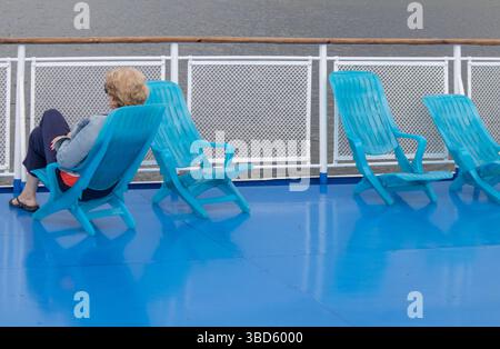 Una donna di mezza età solitaria siede in una chaise longue sul ponte di una nave da crociera, affacciata sul mare. Faccia irriconoscibile. Foto Stock
