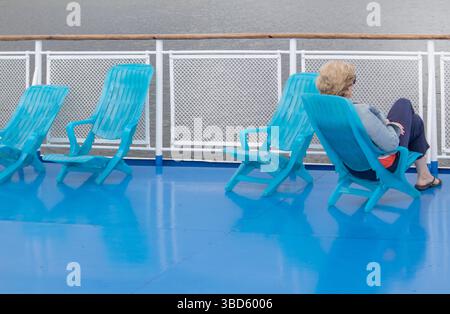 Una donna di mezza età solitaria siede in una chaise longue sul ponte di una nave da crociera, affacciata sul mare. Faccia irriconoscibile. Foto Stock