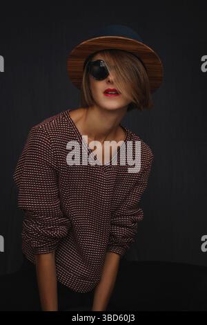 Ritratto della modella su sfondo nero con cappello, occhiali da sole, labbra rosse e maglione. Bella donna con capelli metà che coprono il viso Foto Stock