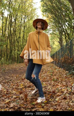 Bella giovane donna che indossa un cappello e un poncho alla moda camminando e divertendosi nella foresta dai colori autunnali. Concetto autunnale Foto Stock