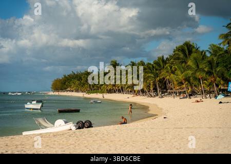 Le Morne, Mauritius - 12 maggio 2024: Persone che camminano e si rilassano su una lunga spiaggia sabbiosa costeggiata da palme e piccole barche ancorate al largo Foto Stock