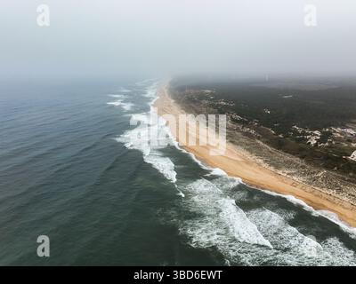 Ripresa aerea di una lunga spiaggia di sabbia che sfuma nella nebbia, sostenuta da foreste e strutture sparse Foto Stock