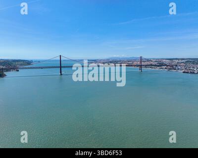 Vista dal drone del Ponte del 25 aprile che si estende sull'acqua con Lisbona spalancata lungo la riva lontana Foto Stock