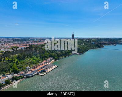 Almada, Portogallo - 12 maggio 2024: Veduta aerea degli edifici abbandonati lungo l'acqua con la statua del Cristo Re in piedi sopra una collina boscosa Foto Stock