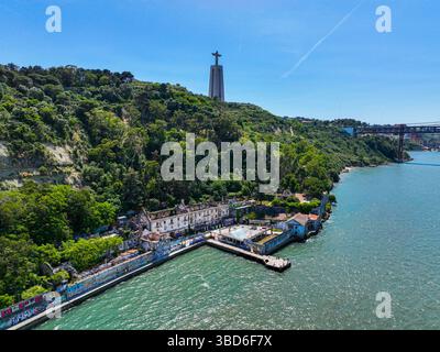 Almada, Portogallo - 12 maggio 2024: Veduta aerea degli edifici abbandonati lungo l'acqua con la statua del Cristo Re in piedi sopra una collina boscosa Foto Stock