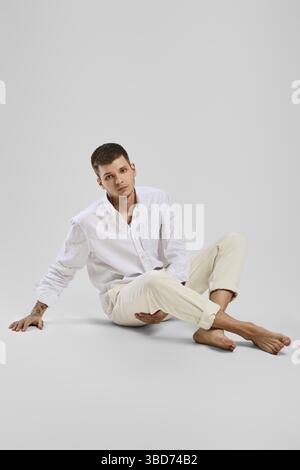 Ritratto studio completo di giovane uomo a piedi nudi in camicia bianca e pantaloni avorio seduti sul pavimento Foto Stock