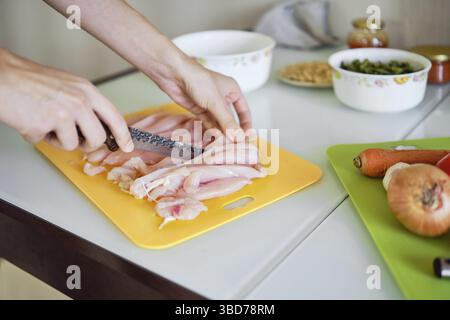 Una persona affetta sapientemente filetti di pollo freschi su un tagliere luminoso con verdure e condimenti disposti nelle vicinanze, mostrando un invito Foto Stock