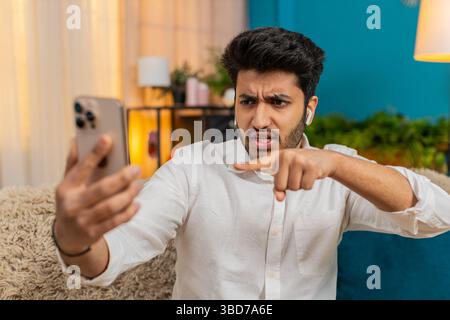 Un uomo indiano a casa sul divano che discute durante una videochiamata, parlando attraverso le cuffie. Ragazzo arabo frustrato, emotivo, alzare la voce, lo stress, reagire intensamente, esprimere disaccordo e tensione Foto Stock