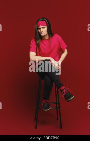 Il giovane uomo adulto si siede sullo sgabello alto con la gamba su un'altra gamba e si appoggia in avanti Foto Stock