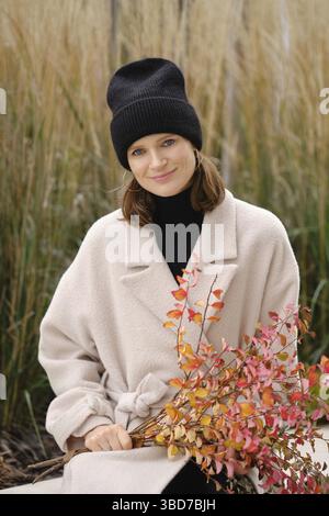 Una donna allegra siede pacificamente circondata da alte erbe, indossando un morbido cappotto beige e un berretto nero Foto Stock