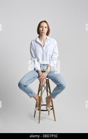 Ritratto di stile di vita di donna di mezza età caucasica a testa rossa in camicia bianca e jeans blu seduti su uno sgabello alto Foto Stock