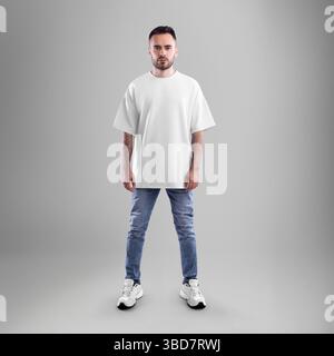 Mockup di t-shirt oversize testurizzata bianca su uomo con barba in jeans blu, sneaker, vista frontale, isolata sullo sfondo. Modello camicia maschio per design, Foto Stock